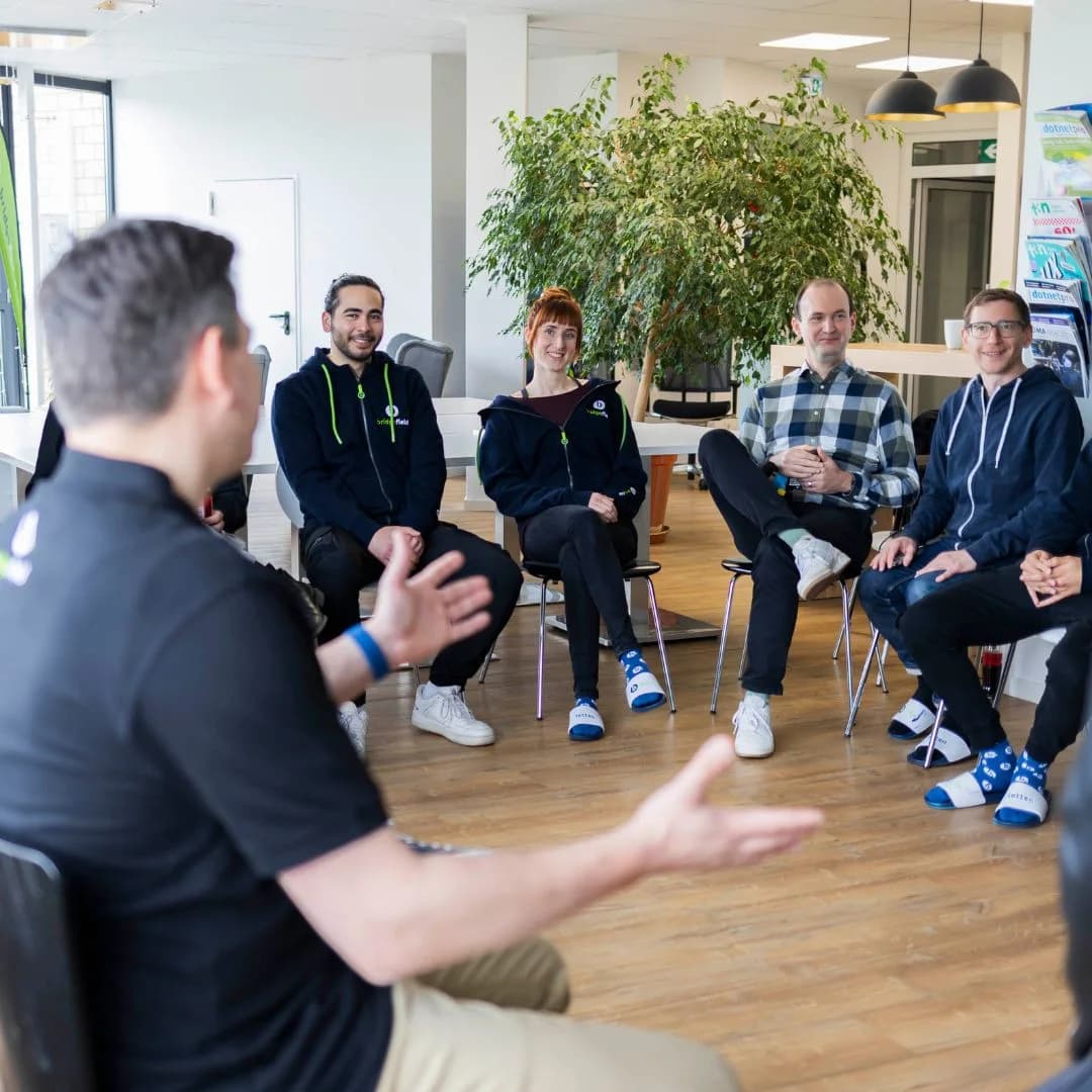 Das Team sitzt im Stuhlkreis, ein Kollege erklärt etwas mit den Händen
